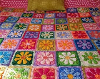 Puede incluir: Una colorida manta de ganchillo hecha a mano con un diseño floral. La manta presenta bloques cuadrados con flores vibrantes en varios colores, incluyendo rosa, azul, amarillo y verde. La manta está colocada sobre una cama con una colcha rosa de lunares y un cojín verde.