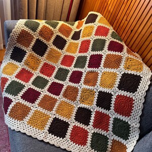 Pu&ograve; includere: Una coperta all'uncinetto fatta a mano con un design patchwork. La coperta presenta quadrati in vari colori, tra cui giallo senape, bordeaux, verde oliva e marrone, con un bordo color crema. La coperta &egrave; drappeggiata su una poltrona grigia.