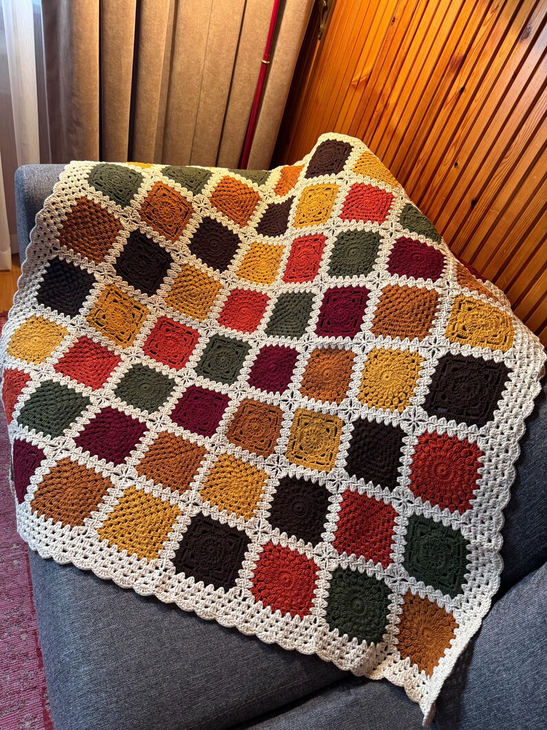 Pu&ograve; includere: Una coperta all'uncinetto fatta a mano con un design patchwork. La coperta presenta quadrati in vari colori, tra cui giallo senape, bordeaux, verde oliva e marrone, con un bordo color crema. La coperta &egrave; drappeggiata su una poltrona grigia.