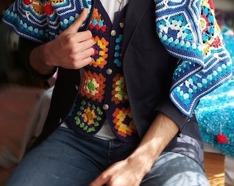 Op de afbeelding: Een persoon draagt ​​een blauwe blazer met een kleurrijk gebreid vest en een blauwe en witte gebreide sjaal. De persoon draagt ​​een blauwe jeans en een wit T-shirt.