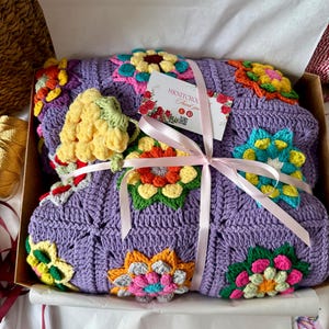 Puede incluir: Una manta de ganchillo lila con vibrantes dise&ntilde;os florales multicolores, presentada en una caja de cart&oacute;n y atada con una cinta rosa. La manta presenta flores amarillas, naranjas, rosas y azules. Se adjunta una peque&ntilde;a etiqueta con texto.