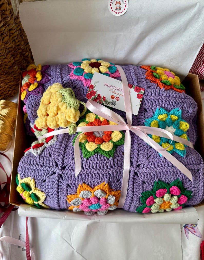 Puede incluir: Una manta de ganchillo lila con vibrantes dise&ntilde;os florales multicolores, presentada en una caja de cart&oacute;n y atada con una cinta rosa. La manta presenta flores amarillas, naranjas, rosas y azules. Se adjunta una peque&ntilde;a etiqueta con texto.