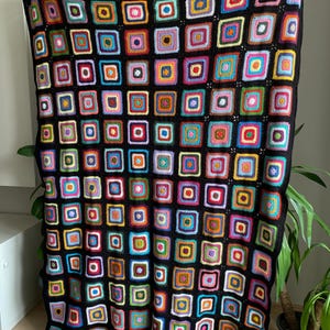 Könnte beinhalten: Eine schwarze Häkeldecke mit einem bunten quadratischen Muster. Die Quadrate sind aus verschiedenen farbigen Garnen gefertigt und in einem Gittermuster angeordnet.