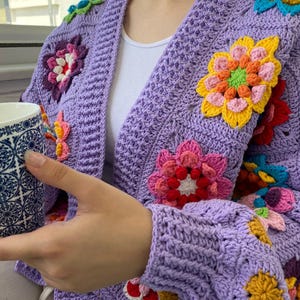 Puede incluir: Un c&aacute;rdigan de ganchillo color lavanda con un patr&oacute;n de puntos texturizados y grandes motivos florales multicolores en tonos amarillo, rosa, rojo y azul. La prenda tiene mangas largas y un dise&ntilde;o abierto, sobre una camiseta blanca.