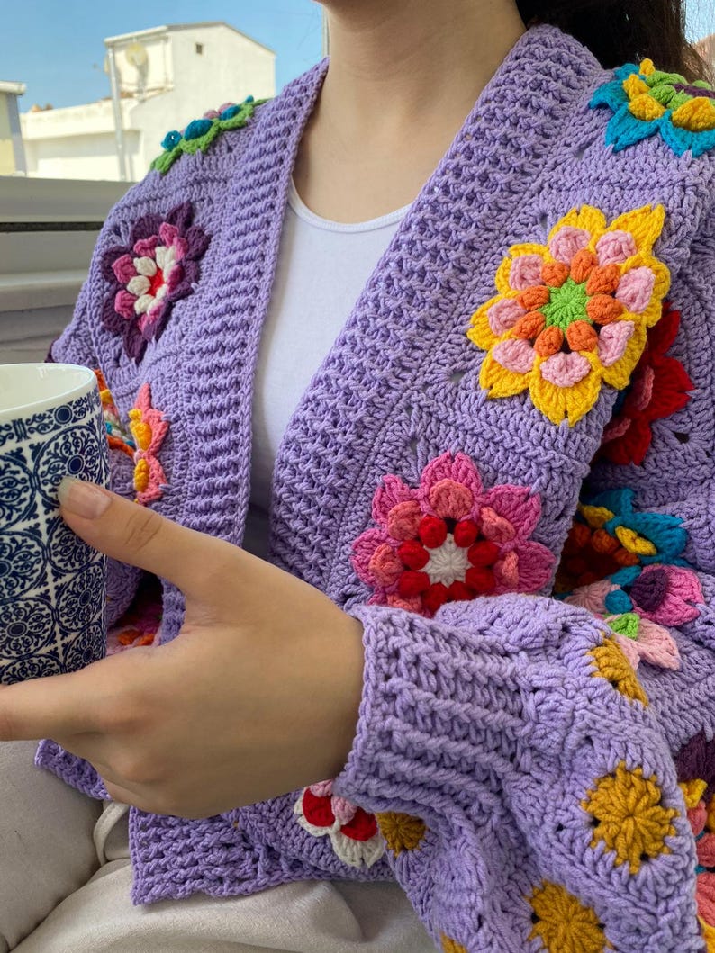 Puede incluir: Un c&aacute;rdigan de ganchillo color lavanda con un patr&oacute;n de puntos texturizados y grandes motivos florales multicolores en tonos amarillo, rosa, rojo y azul. La prenda tiene mangas largas y un dise&ntilde;o abierto, sobre una camiseta blanca.