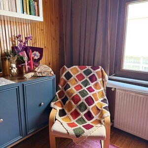 Pu&ograve; includere: Una coperta all'uncinetto fatta a mano e colorata, drappeggiata su una sedia di legno. La coperta presenta un design patchwork con quadrati rossi, arancioni, gialli, verdi e bordeaux. La sedia &egrave; in una stanza con pannelli di legno e una finestra.