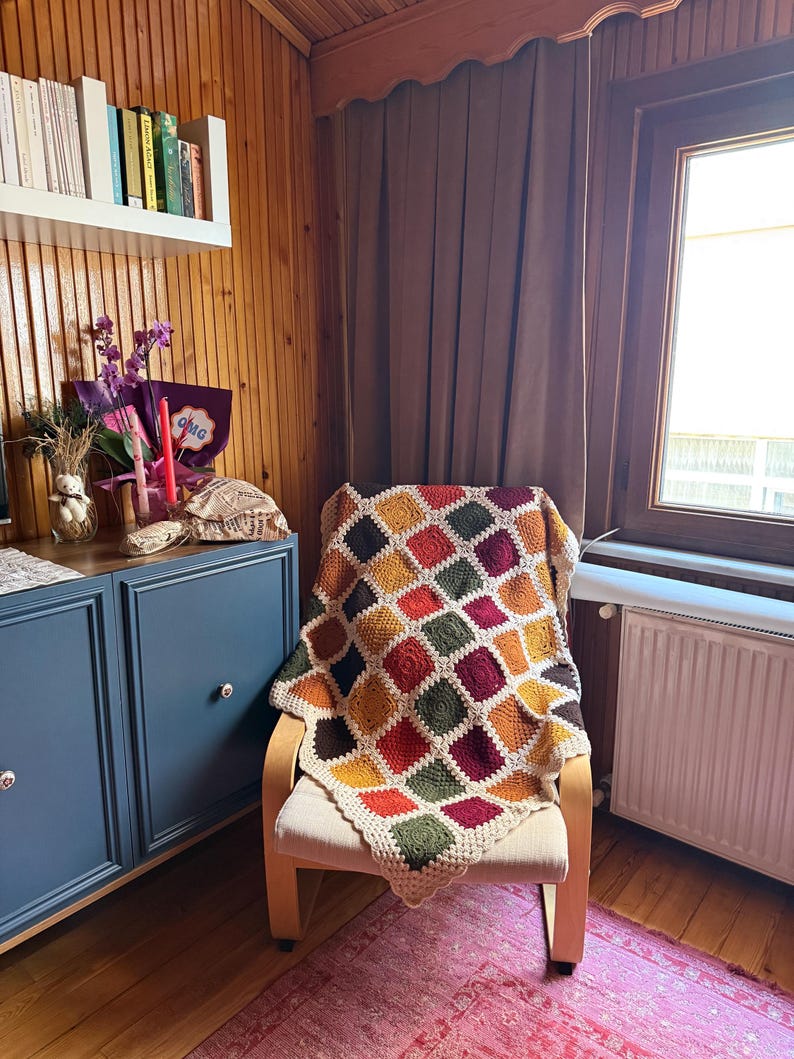 Pu&ograve; includere: Una coperta all'uncinetto fatta a mano e colorata, drappeggiata su una sedia di legno. La coperta presenta un design patchwork con quadrati rossi, arancioni, gialli, verdi e bordeaux. La sedia &egrave; in una stanza con pannelli di legno e una finestra.