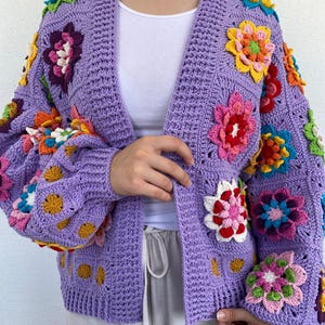 Puede incluir: Una chaqueta de punto de ganchillo de color morado con detalles de flores de colores. La chaqueta tiene cuello en V y mangas largas.