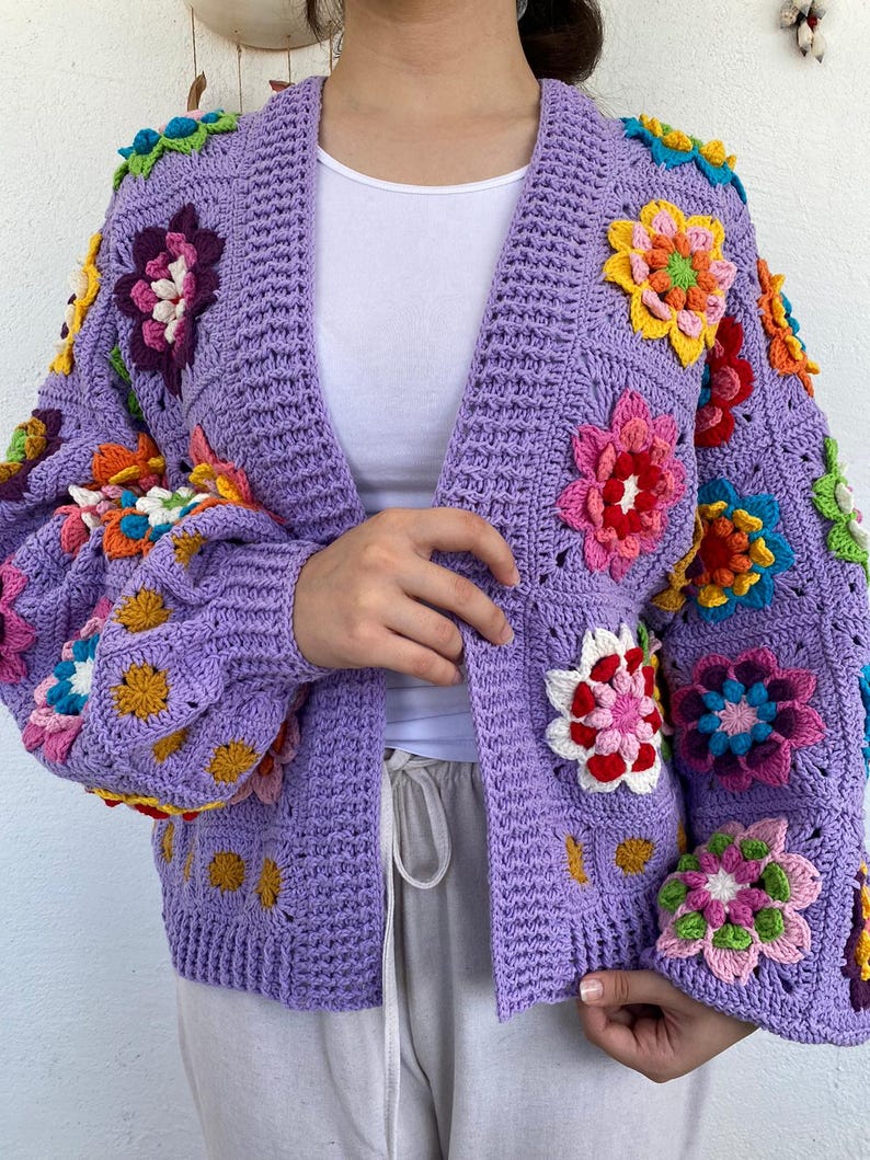 Puede incluir: Una chaqueta de punto de ganchillo de color morado con detalles de flores de colores. La chaqueta tiene cuello en V y mangas largas.