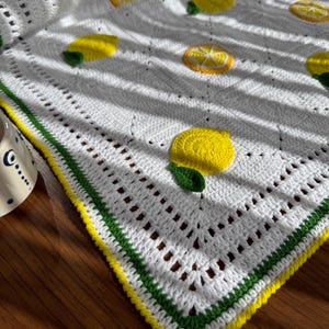 Puede incluir: Una manta blanca de ganchillo con aplicaciones de limones amarillos y hojas verdes. La manta tiene un borde decorativo con rayas amarillas y verdes. La manta está sobre una superficie de madera.