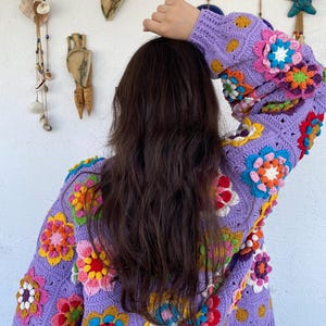 Puede incluir: Un c&aacute;rdigan de crochet hecho a mano en un suave tono lavanda. El su&eacute;ter est&aacute; adornado con coloridas aplicaciones florales tridimensionales en tonos de rosa, amarillo, naranja y verde. El dise&ntilde;o presenta un patr&oacute;n cl&aacute;sico de granny square, creando una prenda acogedora y art&iacute;stica.