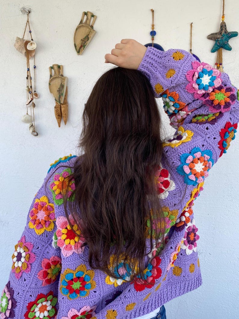 Puede incluir: Un c&aacute;rdigan de crochet hecho a mano en un suave tono lavanda. El su&eacute;ter est&aacute; adornado con coloridas aplicaciones florales tridimensionales en tonos de rosa, amarillo, naranja y verde. El dise&ntilde;o presenta un patr&oacute;n cl&aacute;sico de granny square, creando una prenda acogedora y art&iacute;stica.