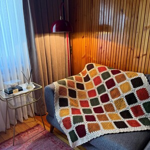 Pu&ograve; includere: Una coperta all'uncinetto fatta a mano con un motivo geometrico di quadrati nei toni dell'arancione, giallo, verde e bordeaux. La coperta &egrave; drappeggiata su un divano grigio, con un bordo bianco. Un elemento di arredo per la casa.