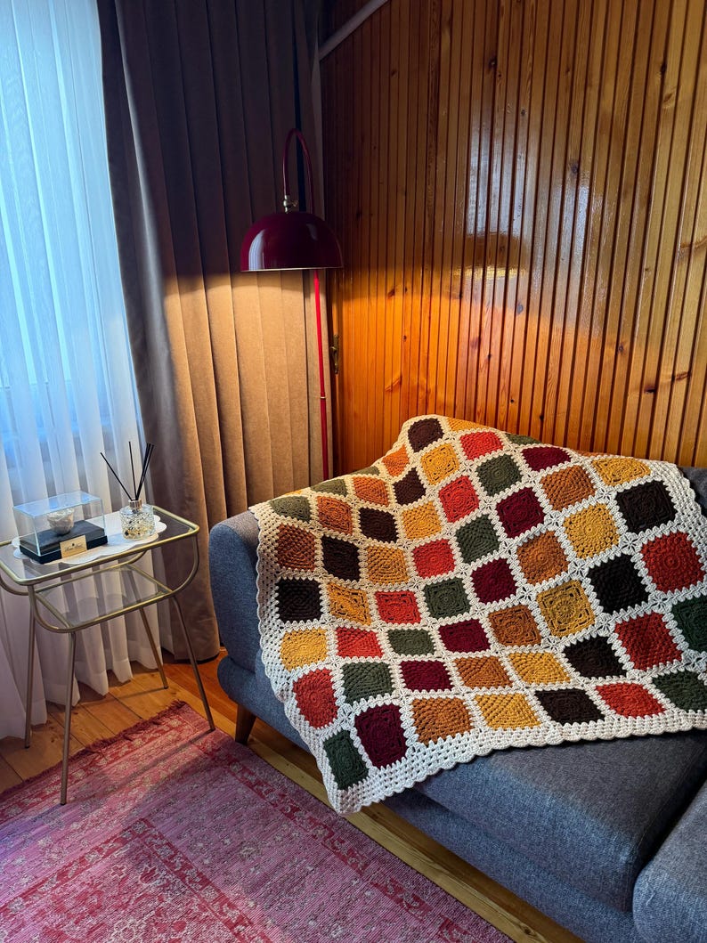 Pu&ograve; includere: Una coperta all'uncinetto fatta a mano con un motivo geometrico di quadrati nei toni dell'arancione, giallo, verde e bordeaux. La coperta &egrave; drappeggiata su un divano grigio, con un bordo bianco. Un elemento di arredo per la casa.