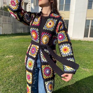 May include: A long black crochet cardigan with a colorful granny square pattern. The cardigan has a tie belt and long sleeves.