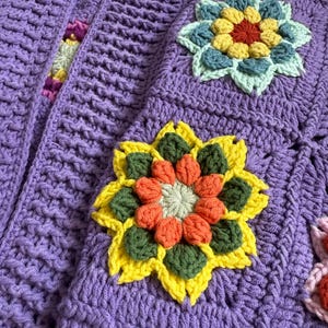 Puede incluir: Primer plano de una manta de ganchillo morada hecha a mano con grandes aplicaciones de flores coloridas. Las flores presentan p&eacute;talos amarillos, naranjas, verdes y azules, a&ntilde;adiendo un contraste vibrante al fondo morado liso. La textura de la manta es visible.