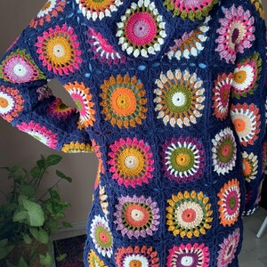 May include: A navy blue crocheted cardigan with a hood, featuring a colorful geometric pattern of circles and squares. The cardigan is made with a variety of yarn colors, including pink, orange, yellow, green, and white.