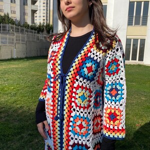 May include: A long, colorful crochet cardigan with a tie closure. The cardigan is made of granny squares in shades of red, orange, yellow, green, blue, and white. The cardigan has a navy blue trim.