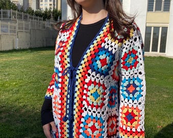 Puede incluir: Una larga chaqueta de ganchillo de colores con cierre de lazo. La chaqueta está hecha de cuadrados de la abuela en tonos de rojo, naranja, amarillo, verde, azul y blanco. La chaqueta tiene un ribete azul marino.