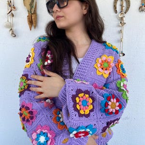 Puede incluir: Una chaqueta de punto de ganchillo de color morado con detalles de flores de colores. La chaqueta tiene cuello en V y mangas largas.