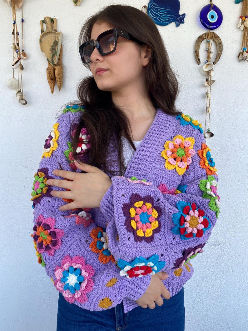 Puede incluir: Una chaqueta de punto de ganchillo de color morado con detalles de flores de colores. La chaqueta tiene cuello en V y mangas largas.
