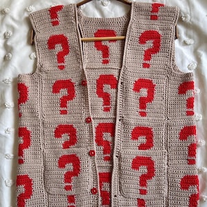 May include: A beige, handmade crochet vest featuring a repeating pattern of red question marks. The vest has a button-up front, two pockets, and is displayed on a wooden hanger. The garment is made of a textured yarn.