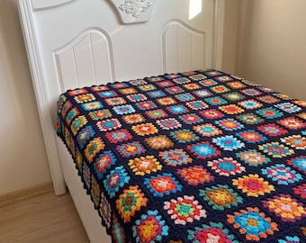 Op de afbeelding: Een kleurrijk granny square gehaakt deken in tinten blauw, rood, oranje, geel, groen en roze op een wit bedframe.