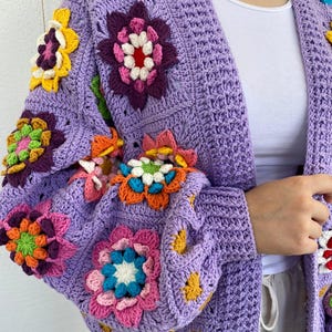 Puede incluir: Una chaqueta de punto de ganchillo de color morado con detalles de flores de colores. La chaqueta tiene cuello en V y mangas largas.