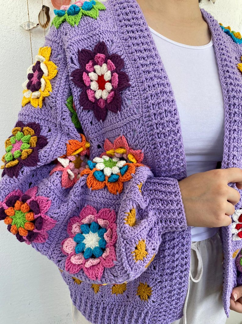 Puede incluir: Una chaqueta de punto de ganchillo de color morado con detalles de flores de colores. La chaqueta tiene cuello en V y mangas largas.