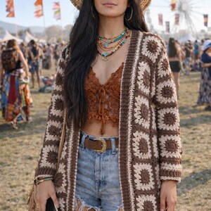 May include: A woman wearing a brown and cream crochet cardigan, brown lace top, denim shorts, and a straw hat. She is also wearing sunglasses, jewelry, and a brown belt. The background shows a crowd of people and a Ferris wheel.