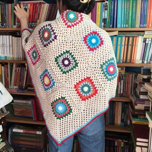 May include: A beige crochet shawl with a colorful geometric pattern. The shawl has a fringe tassel at the bottom and is made with a granny square stitch.