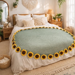 May include: A round, sage green crochet blanket with a sunflower border. The blanket is displayed on a bed with white linens. The sunflowers have yellow petals and brown centers. The room has a bohemian aesthetic.