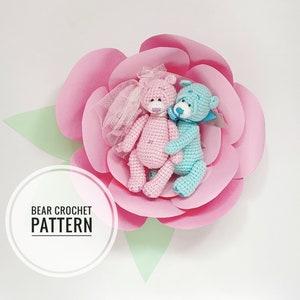 Pode incluir: Dois ursos de crochê, um rosa e outro azul, estão sentados juntos em uma flor de papel rosa. O urso rosa tem um véu branco e o urso azul tem um laço azul. O texto "BEAR CROCHET PATTERN" está escrito em um círculo branco.