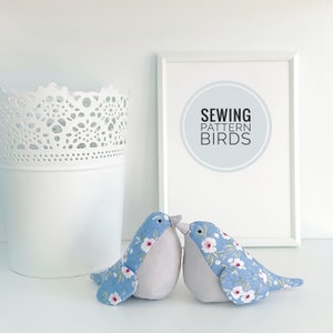 Puede incluir: Dos pájaros de tela azul y gris con patrones florales se besan. Los pájaros están sentados sobre una superficie blanca. Un marco blanco con el texto "Sewing Pattern Birds" está en el fondo.