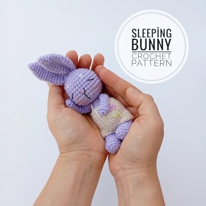 May include: A crocheted purple sleeping bunny with a beige dress and embroidered flowers. The bunny is held in the palms of two hands. The text "SLEEPING BUNNY CROCHET PATTERN" is in a white circle.