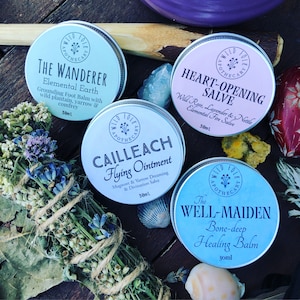 May include: Four small silver tins with labels and lids. The labels are white with black text. The tins contain natural remedies. The labels read "The Wanderer Elemental Earth Grounding Foot Balm", "Heart-Opening Salve Wild Rose, Lavender & Nettle Elemental Fire Salve", "Cailleach Flying Ointment Mugwort & Yarrow Dreaming & Divination Salve", and "The Well-Maiden Bone-deep Healing Balm".
