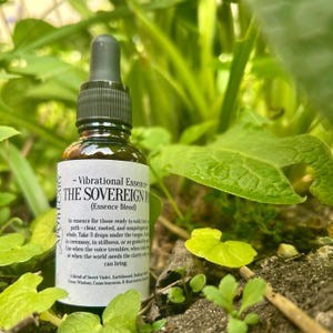 以下が含まれることがあります： ドロップトップ付きの茶色のガラス瓶で、「Vibrational Essence - The Sovereign Path (Essence Blend)」とラベル付けされています。瓶は緑の葉と茶色の土のベッドの上に置かれています。