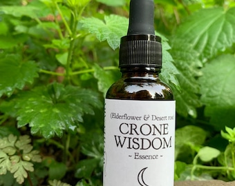 Puede incluir: Una botella de vidrio marrón oscuro con un gotero negro. La botella está etiquetada con "Crone Wisdom - Essence - (Elderflower & Desert Rose)" y presenta un símbolo de luna creciente. La etiqueta también incluye el texto "Folk Magic, Wild Medicine, Natural Herbs" y la dirección del sitio web "www.wildfolkapothecary.com".