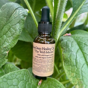 Puede incluir: Una botella de vidrio marrón de "One-Deep Healing Oil" con un cuentagotas negro, sobre un fondo de hojas verdes. La etiqueta incluye texto sobre los ingredientes y beneficios del aceite. El producto es para el cuidado de la piel.