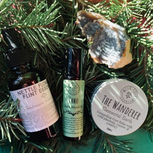 May include: Three natural wellness products: a brown glass bottle with a white label that reads "Nettle Flower Flint Essence Transformation - Car", a small brown glass roller bottle with a white label that reads "Danu Earth Ritual Oil", and a white jar with a black label that reads "The Wanderer Elemental Earth Grounding Foot Balm with wild plantain, yarrow & comfrey 30ml".