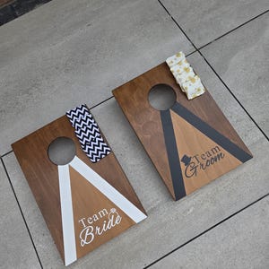 May include: Two wooden cornhole boards with a brown stain, each featuring a circular hole and a design. One board says "Team Bride" with a white and gold design, and the other says "Team Groom" with a black and gold design. Cornhole bean bags are included.