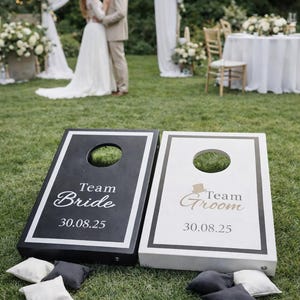 May include: Two cornhole boards, one black and one white, with the words "Team Bride" and "Team Groom" in elegant script. The boards have a hole in the center and are set on a grassy lawn, with bean bags nearby. Wedding theme.