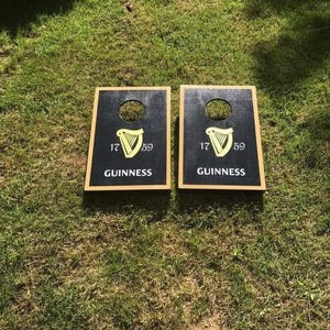 May include: Two black cornhole boards featuring the Guinness logo, a yellow harp, and the text "17 59 GUINNESS". Each board has a hole and a wooden frame. They are placed on a grassy surface.