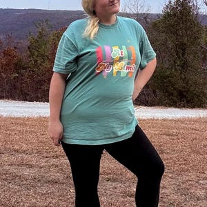Puede incluir: Una camiseta turquesa de manga corta con el texto "At My Limit" en letras coloridas. La camiseta se combina con leggings negros y zapatos negros con suelas blancas. El fondo presenta un paisaje con árboles y un cielo nublado.