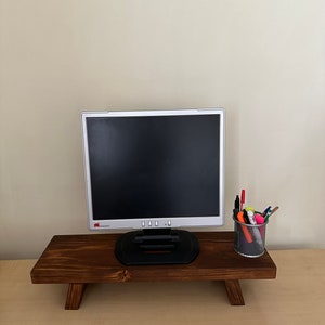May include: A wooden monitor stand with a dark brown stain. A computer monitor sits on top of the stand. A small wire mesh cup with colourful pens and markers sits on the right side of the stand.