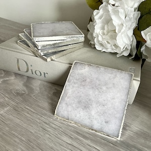 May include: Set of four white agate coasters with silver trim. The coasters are stacked on top of a book with the word "Dior" printed on the cover.