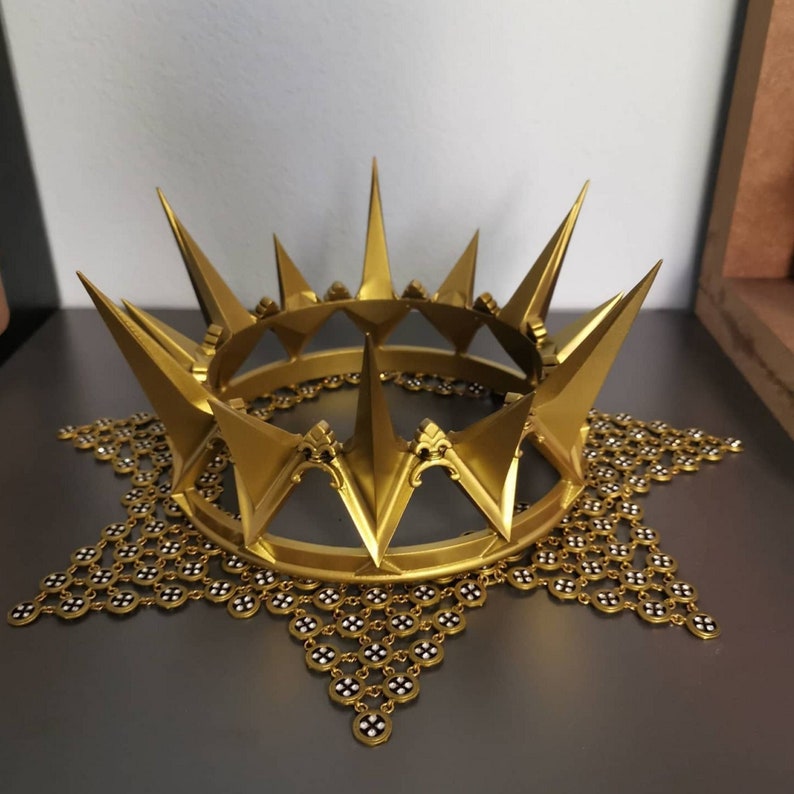 May include: A gold-colored crown with sharp, upward-pointing spikes. The crown sits atop a decorative, star-shaped base with a chain-like pattern. The crown's design features a series of alternating spikes and decorative elements, creating a regal appearance.