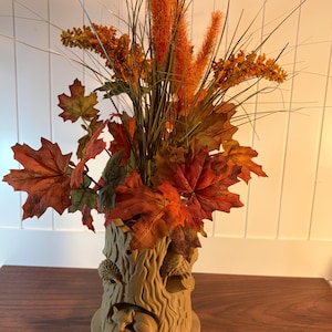 May include: A decorative fall-themed arrangement featuring a beige tree trunk vase with a sleeping fox and other woodland creatures. The vase holds orange and red faux maple leaves, tall orange grass plumes, and other fall foliage.