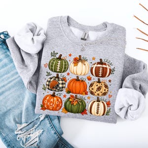 Puede incluir: Una sudadera gris jaspeado de cuello redondo con un estampado gráfico de nueve calabazas decoradas con motivos de fútbol americano, incluyendo cordones, cascos y estrellas, intercaladas con pequeñas hojas naranjas y corazones.