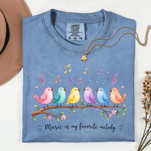 Puede incluir: Una camiseta azul con un gráfico de seis pájaros coloridos posados en una rama marrón. Sobre los pájaros hay varias notas musicales en rosa, amarillo y azul. Debajo de los pájaros, el texto dice: "Music is my favorite melody." Tiene una etiqueta Comfort Colors.